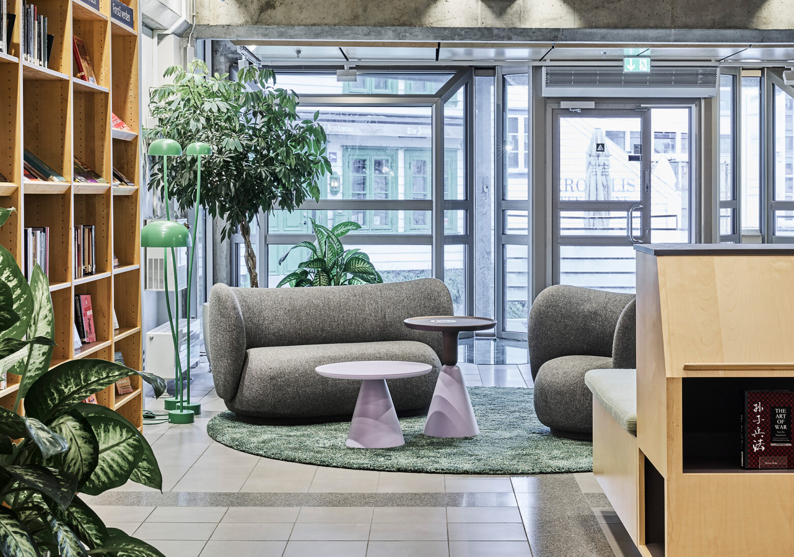 Lesekrok med grå buede sofaer og lilla bord plassert på et rundt grønt teppe foran store vinduer.