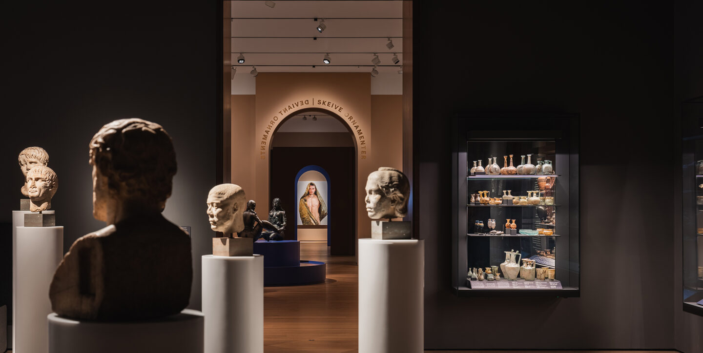 Stemningsbilde fra en mørkere del av galleriet, med byster på hvite søyler som leder øyet mot en opplyst buegang.