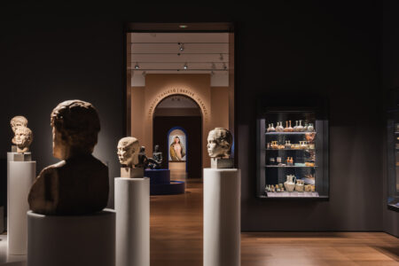 Stemningsbilde fra en mørkere del av galleriet, med byster på hvite søyler som leder øyet mot en opplyst buegang.