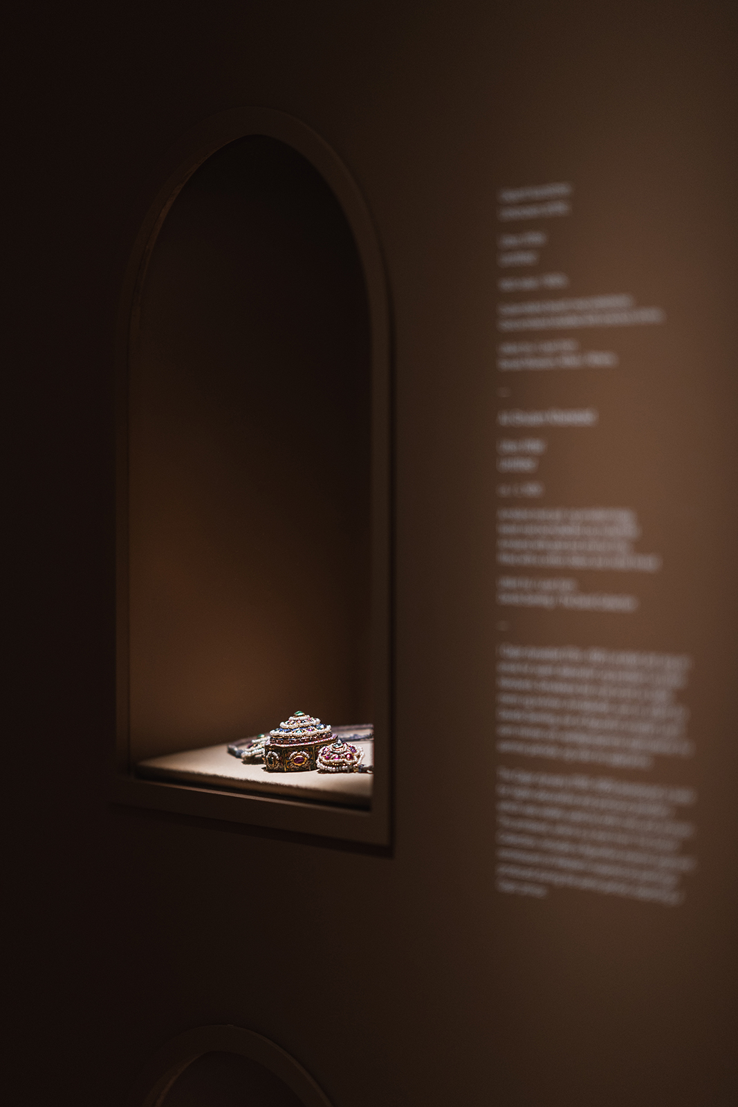 Stemningsfullt bilde av et lite, detaljert smykke utstilt i en belyst, mørk nisje i utstillingsveggen.