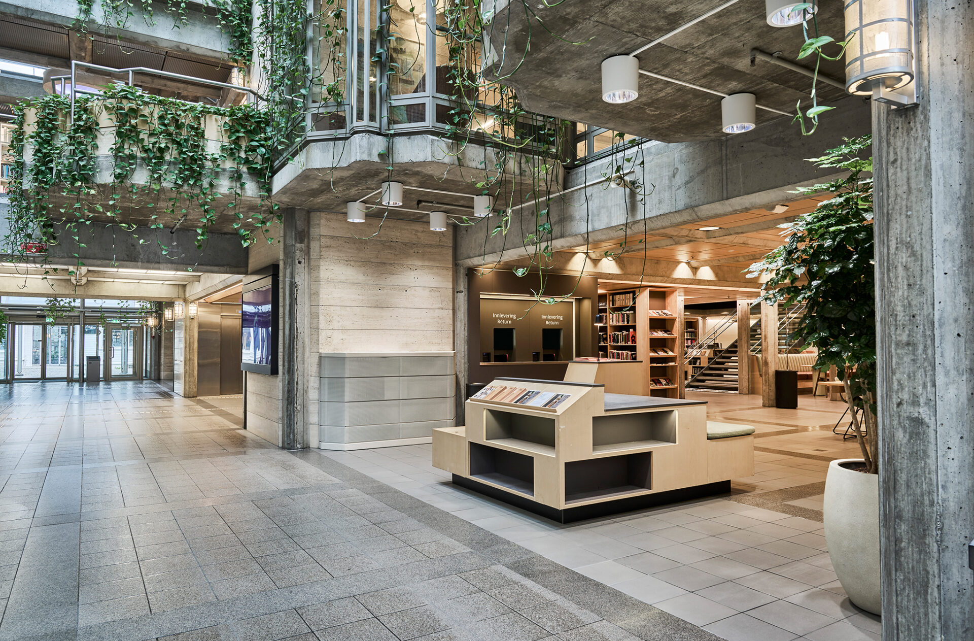 Lysfylt atrium i betong og treverk med hengeplanter, integrert informasjonsskranke og utstillingsmøbler i lys finér.