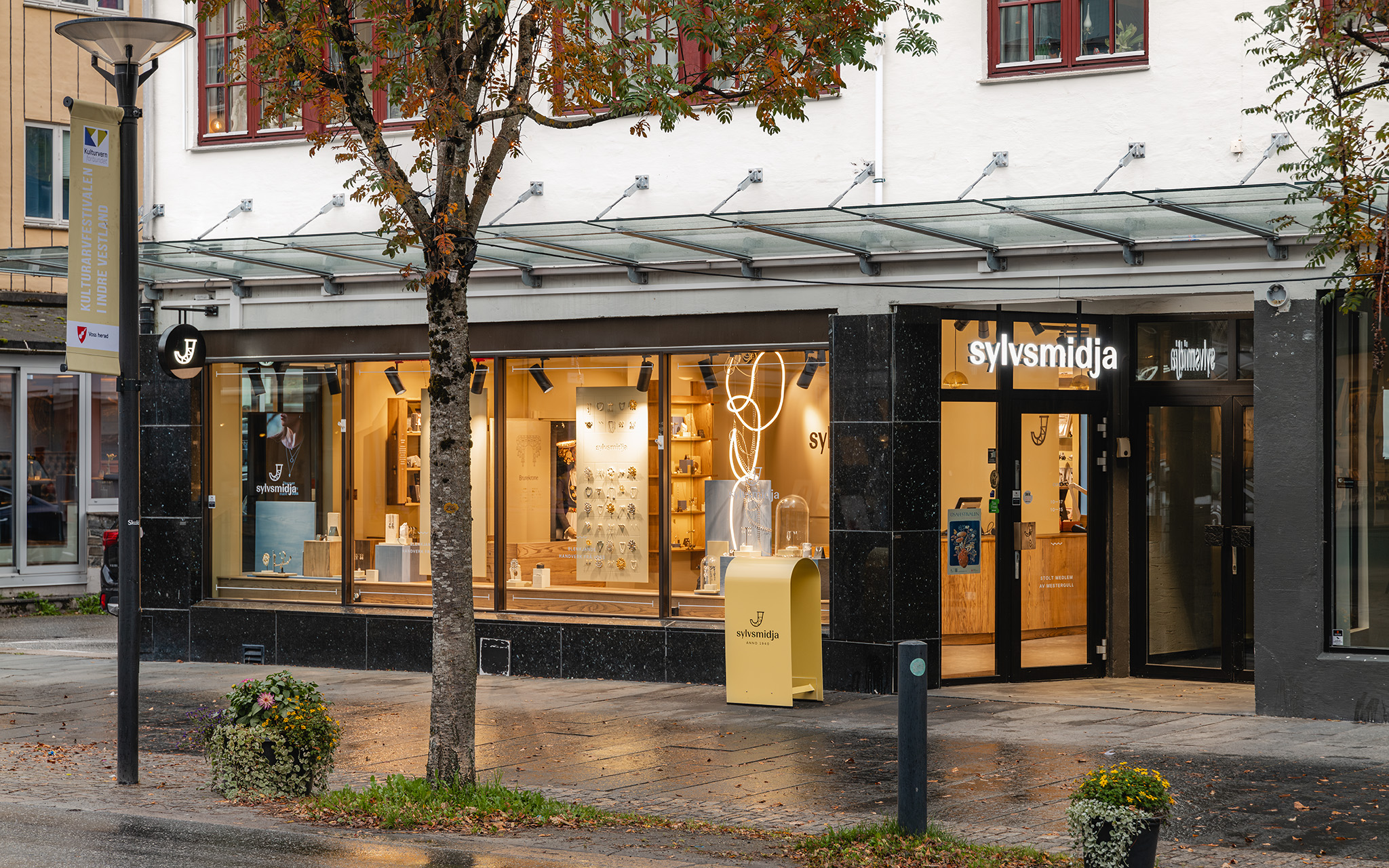 Butikkfasade i mørk poleret stein med hengende glasstak, opplyst vindusfront og frittstående skilt med logo foran inngangen i skumringslys.