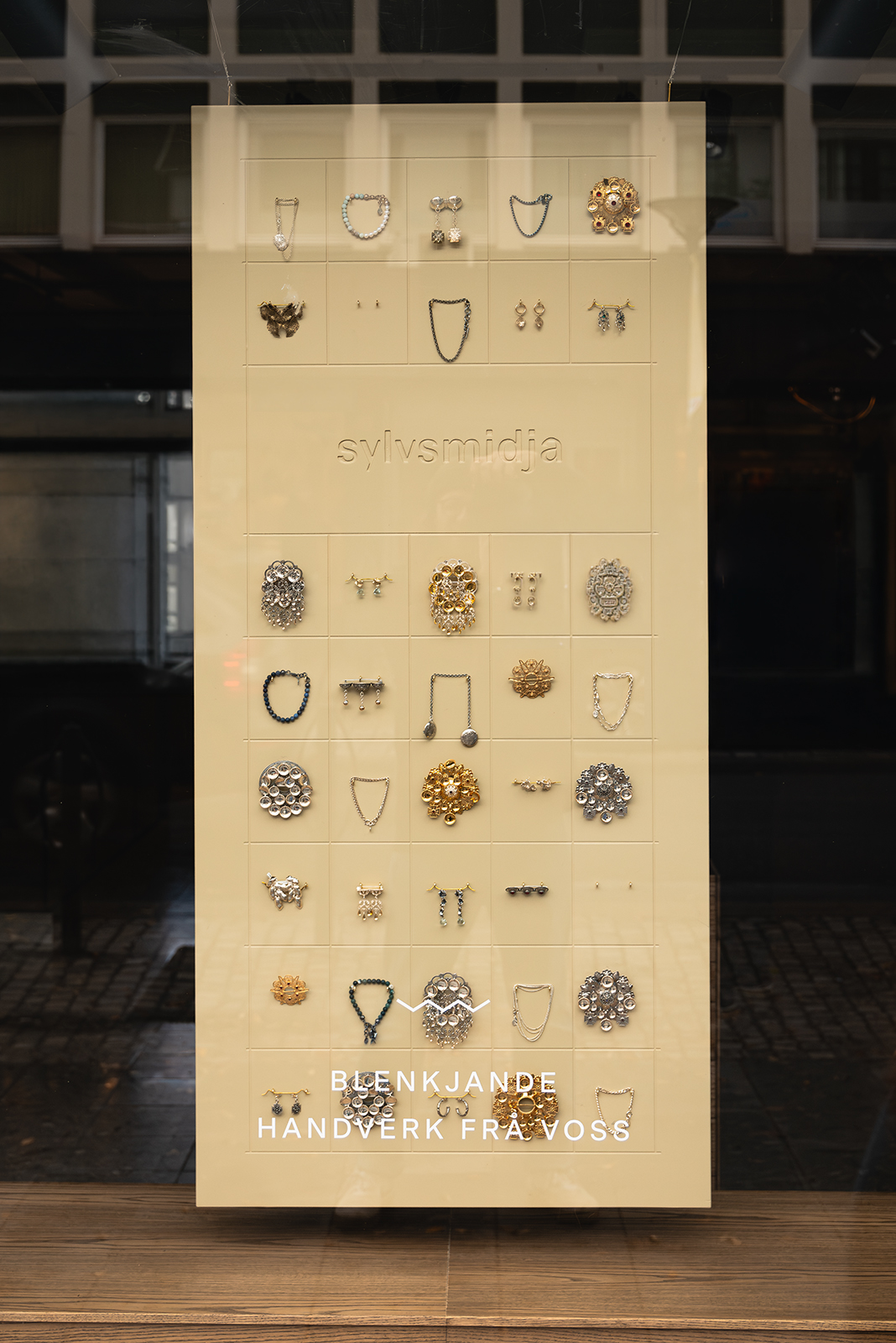Beige panel i vindu satt opp i rutenett med enkeltstående smykker, logo i midten og teksten «Blenkjande handverk frå Voss» nederst.