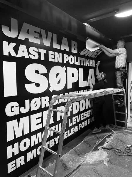To personer monterer stor, hvit folietekst på en svart utstillingsvegg fra et stillas.