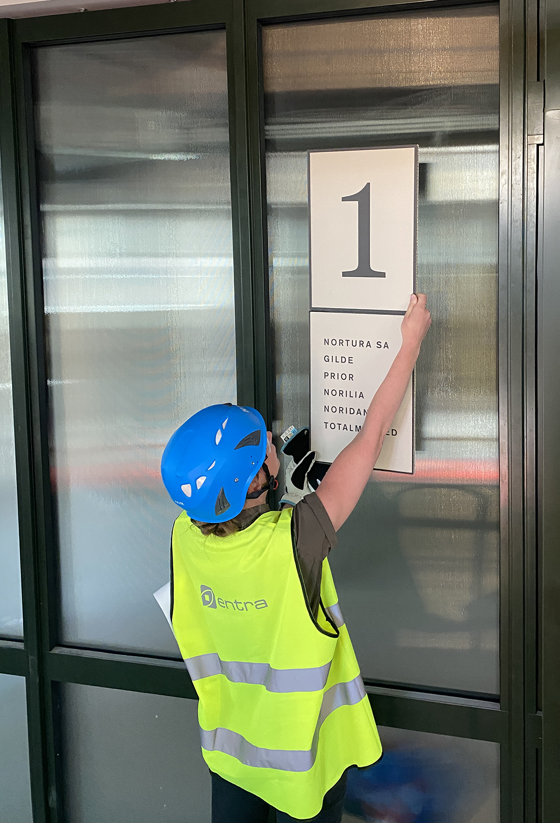 En person i gul refleksvest og vernehjelm vurderer en skiltprototype med tallet 1 og firmanavn på en glassvegg.